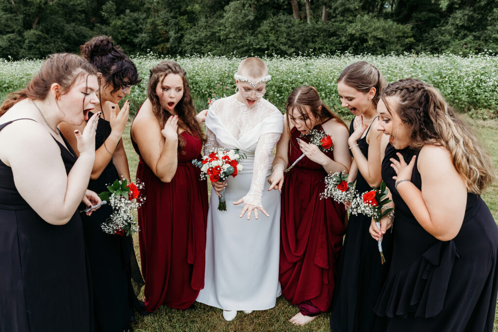 A group of people in formal attire express surprise and admiration at a person's ring, suggesting a marriage proposal or wedding celebration in a grassy field.