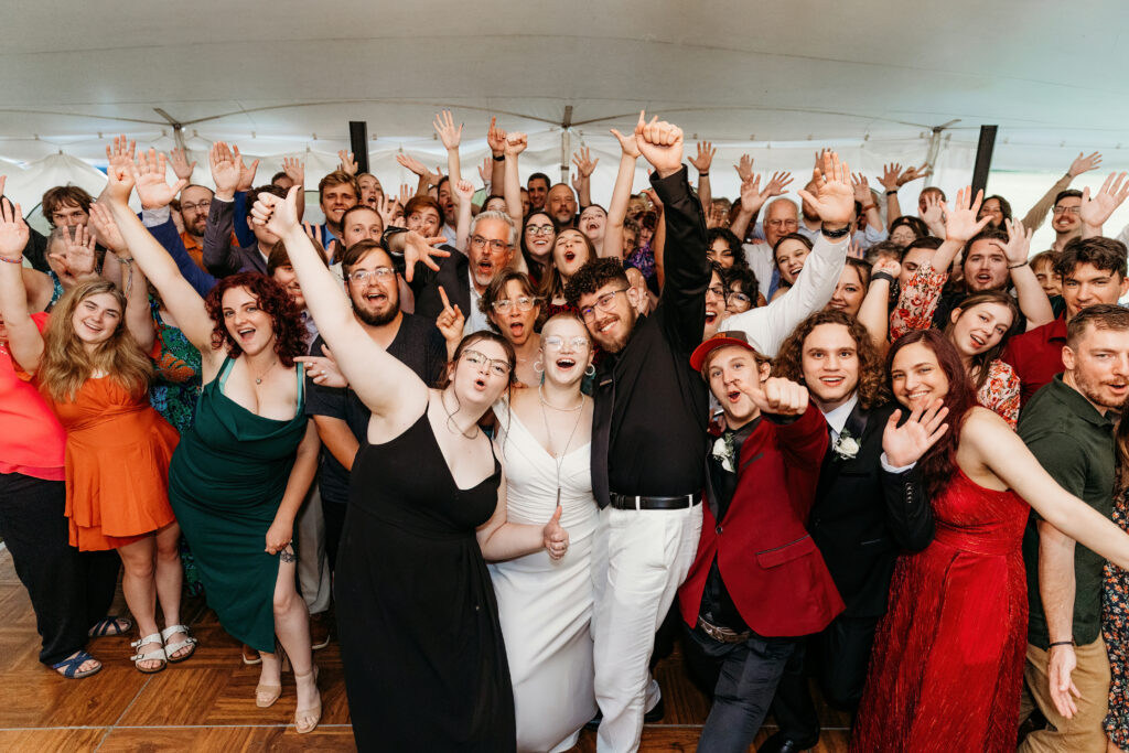 A group of joyful people is celebrating, with many raising their arms and smiling widely in a tented event space. They appear to be having fun.