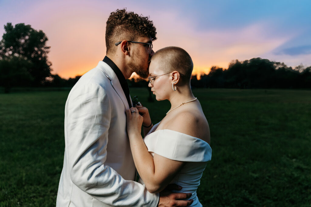 Two people in formal wear embracing outdoors at dusk, with a colorful sunset sky in the background and a green field around them.