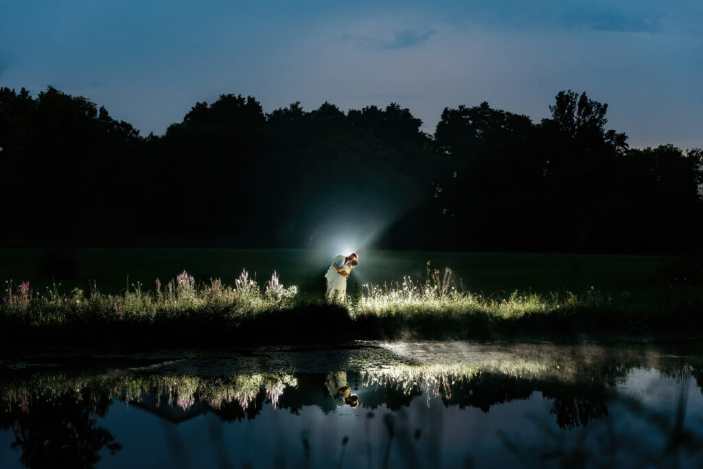 Two people are embracing on a grassy peninsula, backlit by a beam of light at dusk, with their reflection mirrored in calm water.