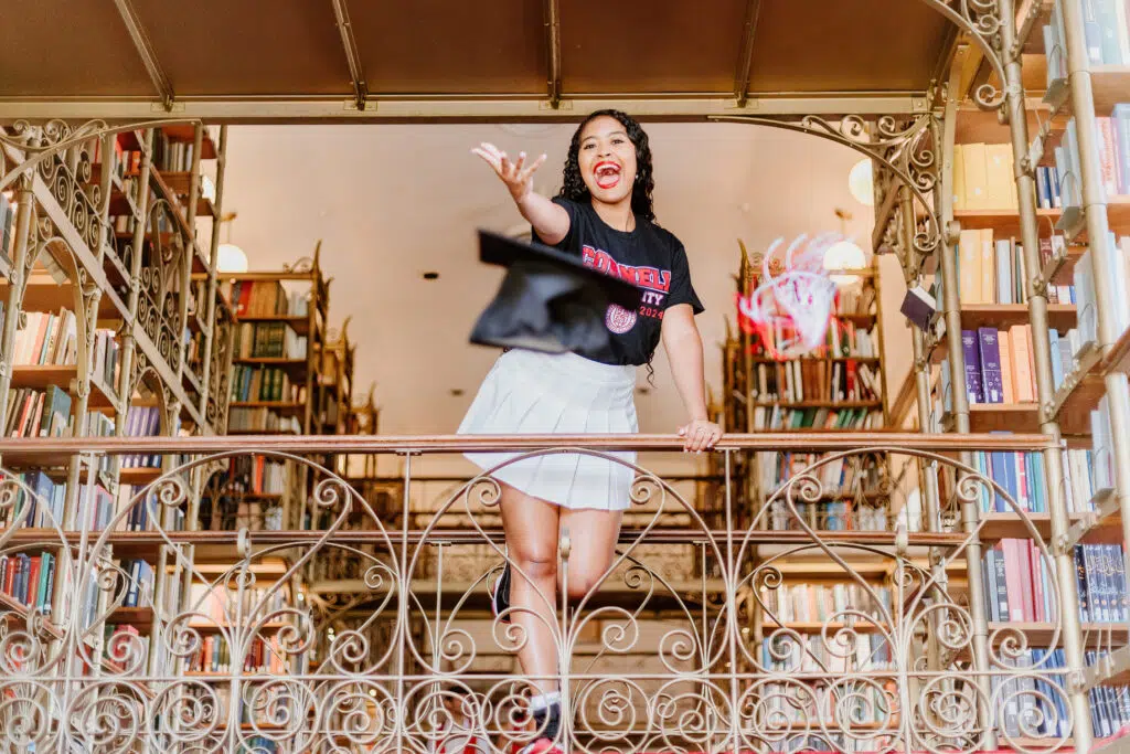 A person is standing on an ornate balcony inside a bookstore, throwing a graduation cap, and celebrating with a big smile. Shelves of books surround them.