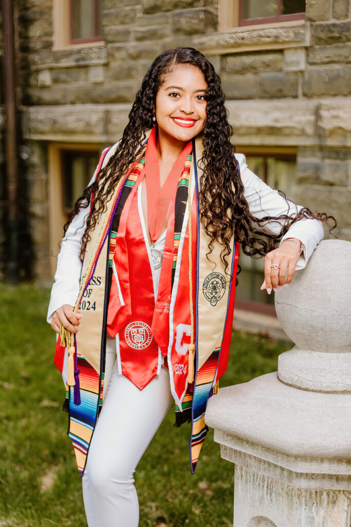 A smiling person wearing a white blouse, white pants, and multiple colorful graduation stoles standing by a stone balustrade outside a building.