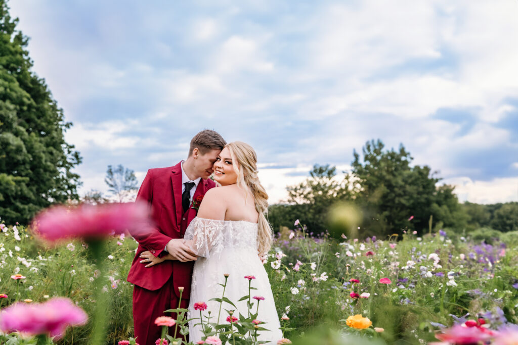 Two people are embracing in a colorful flower field under a cloudy sky; one is wearing a red suit, and the other a white dress.