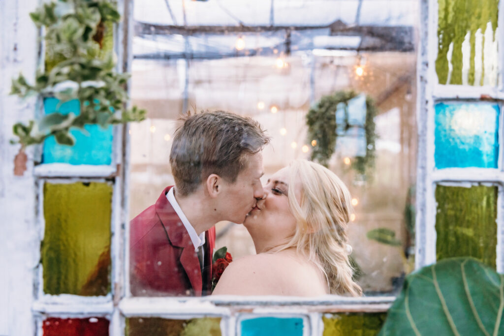 Two people are sharing a kiss, framed by a colorful stained-glass window. There are string lights and greenery in the background, creating a romantic ambiance.