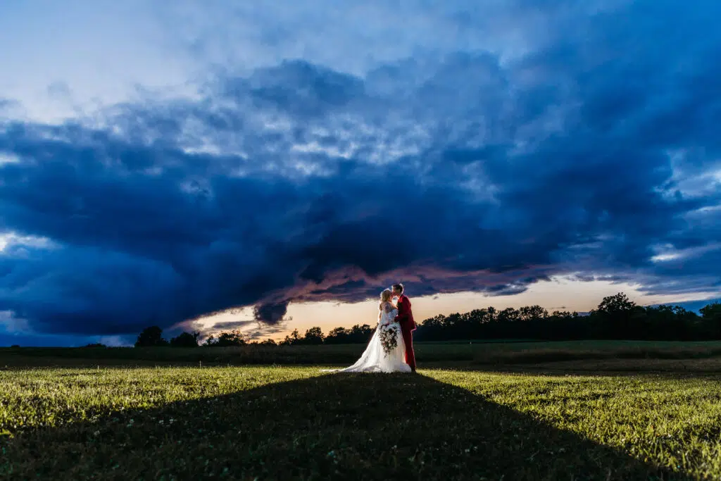 Two people in wedding attire embrace in a field under a dramatic sky with dark clouds and a hint of sunset at the horizon.