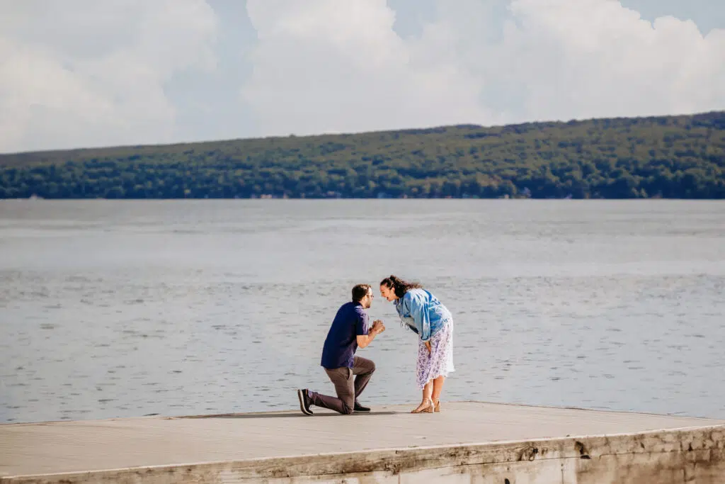 A person is kneeling on one knee facing another person on a lake dock, possibly in a proposal gesture, with hills and a cloudy sky background.