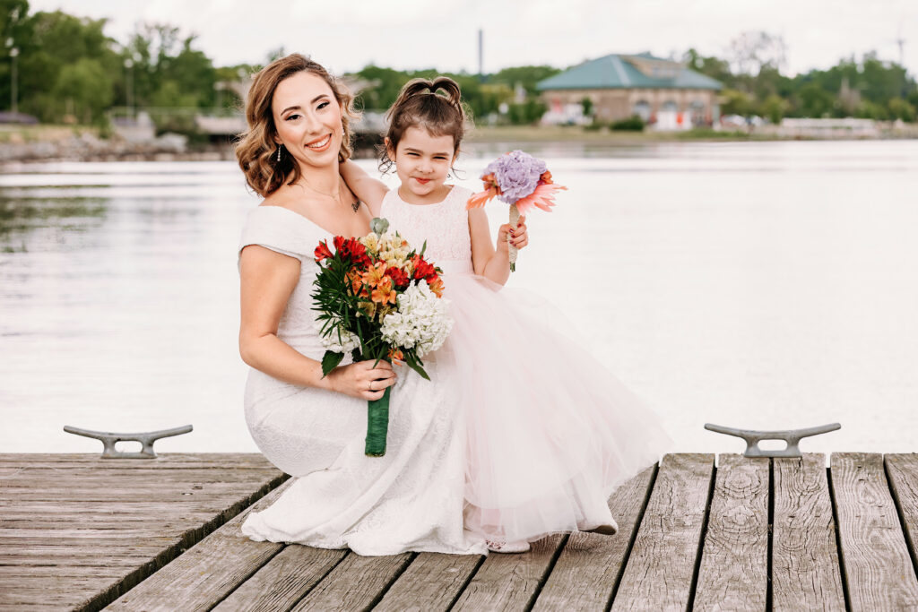 A person in a white dress and a young girl in a tulle skirt hold bouquets on a wooden dock by the water, smiling joyfully.