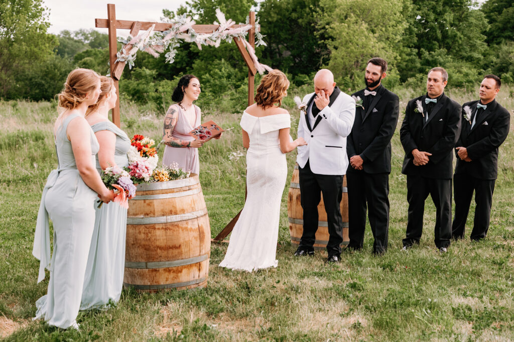 A wedding ceremony outdoors with a person in a white dress, person wiping tears, and attendants wearing gray and black suits beside a floral arch.