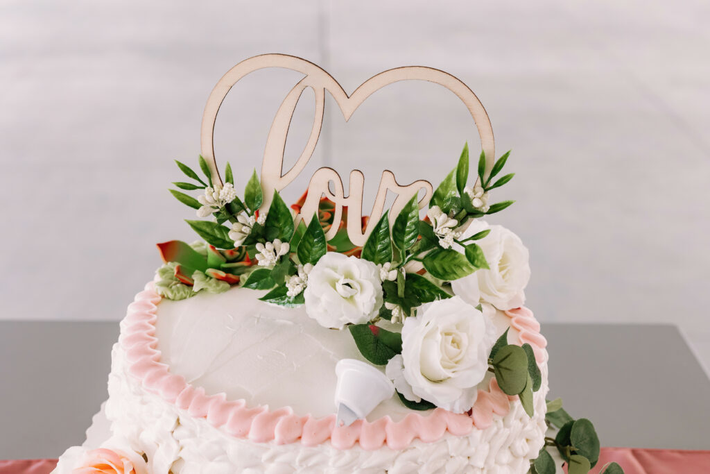 The image shows a cake with pink ruffles, white frosting, and a topper with two hearts and the word