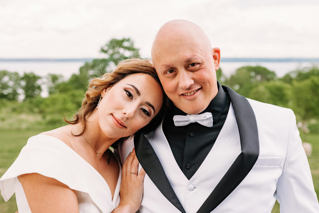 Two people in formal attire smile outdoors, possibly a wedding scene, with trees and a lake in the background. They appear happy and content.
