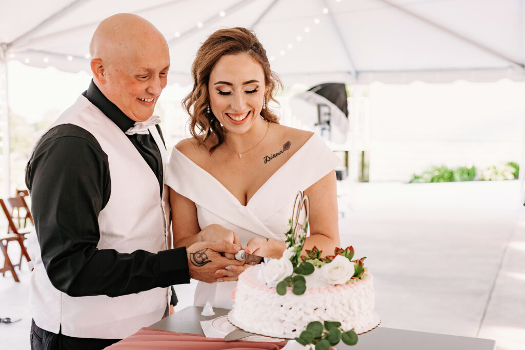 Two people dressed formally, presumably a couple, are happily cutting a white frosted cake adorned with flowers at an event decorated with string lights.
