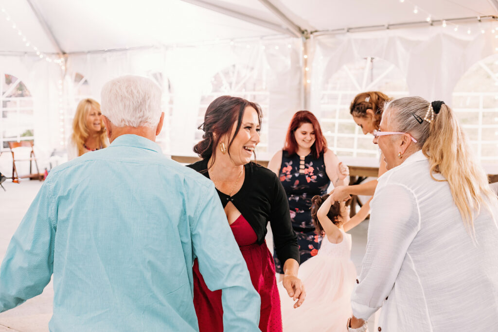 A group of people are happily interacting at an event under a white tent with string lights. A person in a red dress is smiling joyfully.