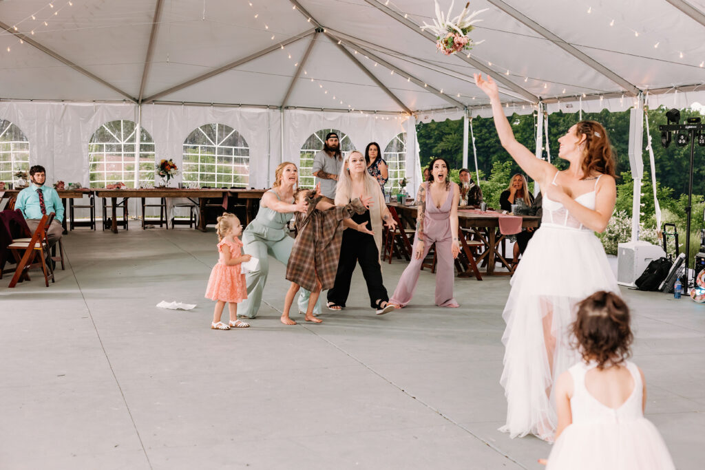 Guests excitedly reach out to catch a bouquet tossed by a bride under a tent at a wedding reception; a moment of joyful anticipation captured.