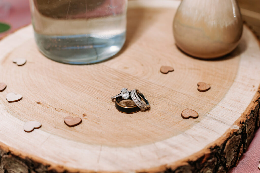 Three rings, possibly wedding bands and an engagement ring, are stacked on a wooden surface with small heart-shaped decorations and vases nearby.