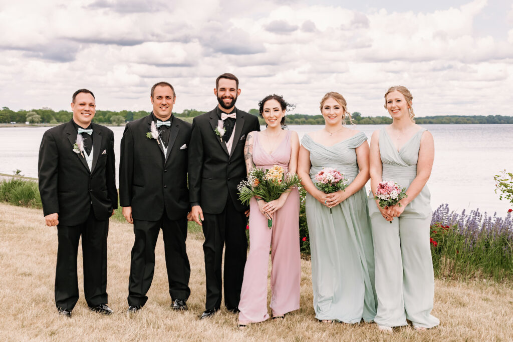 Three people in suits and three people in dresses pose smiling by a lake with cloudy skies, likely a wedding party, with bouquets and boutonnieres.