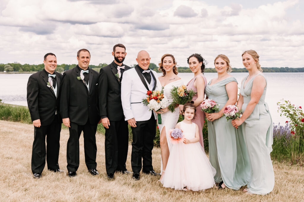 A wedding group photo by a lakeside with seven adults dressed formally and a child holding flowers, under a cloudy sky. They are smiling.
