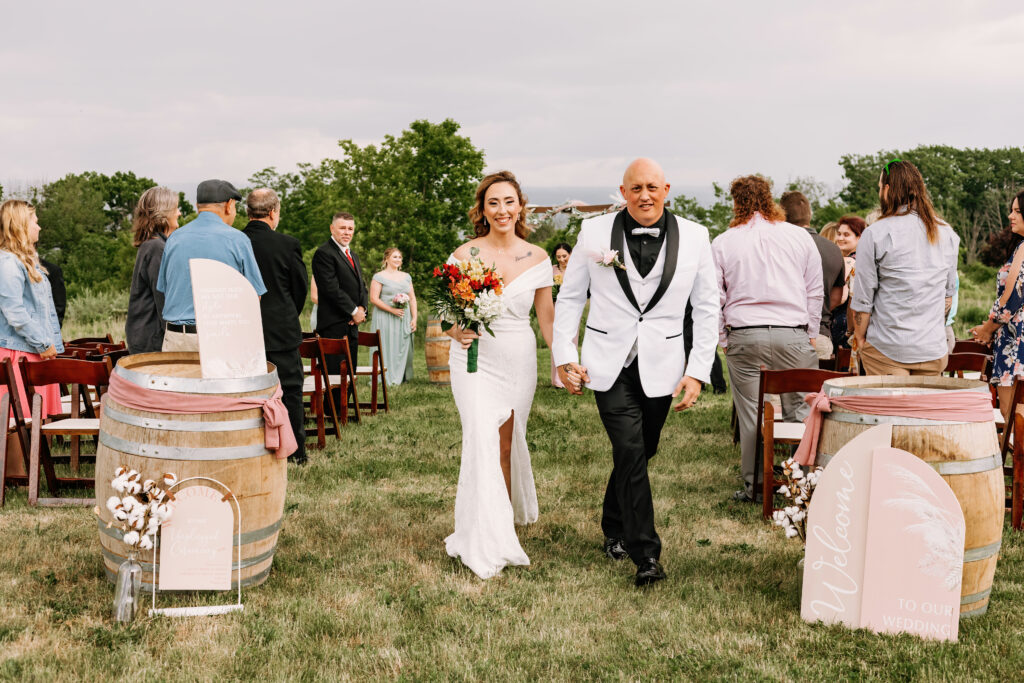 A person in white attire walks down an aisle with a smiling person in a wedding dress, both looking joyful amid guests standing at an outdoor ceremony.