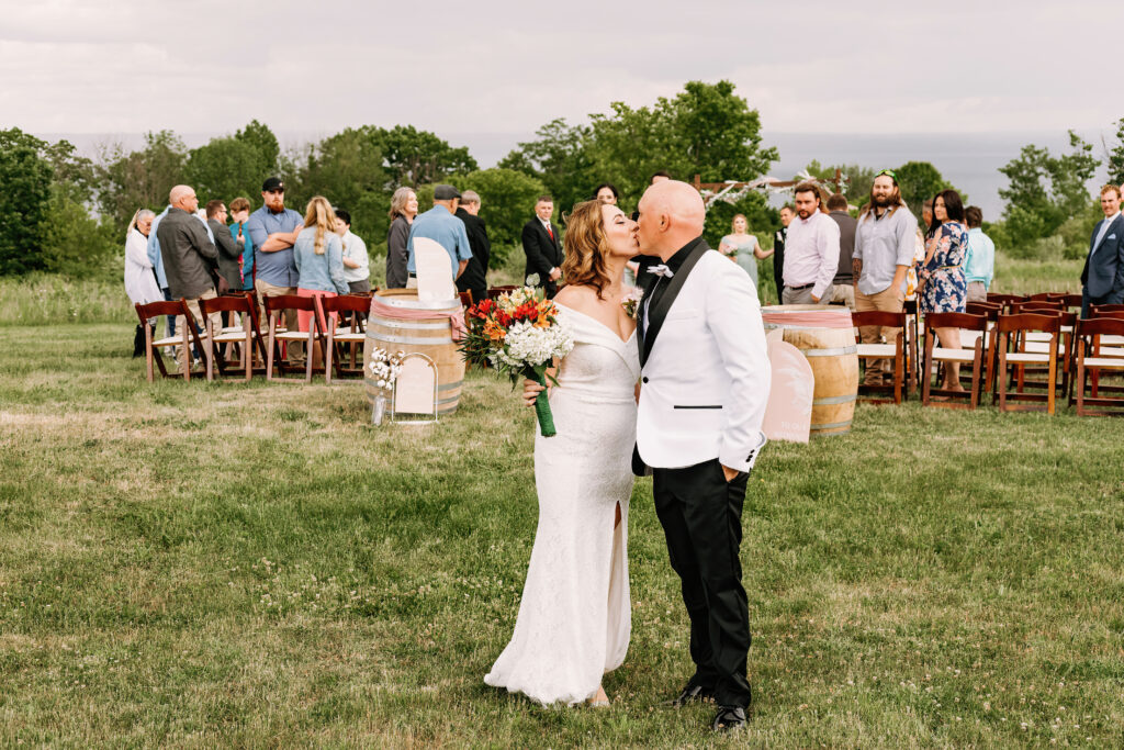A person and a person in wedding attire share a moment among guests at an outdoor ceremony with open field and cloudy sky.