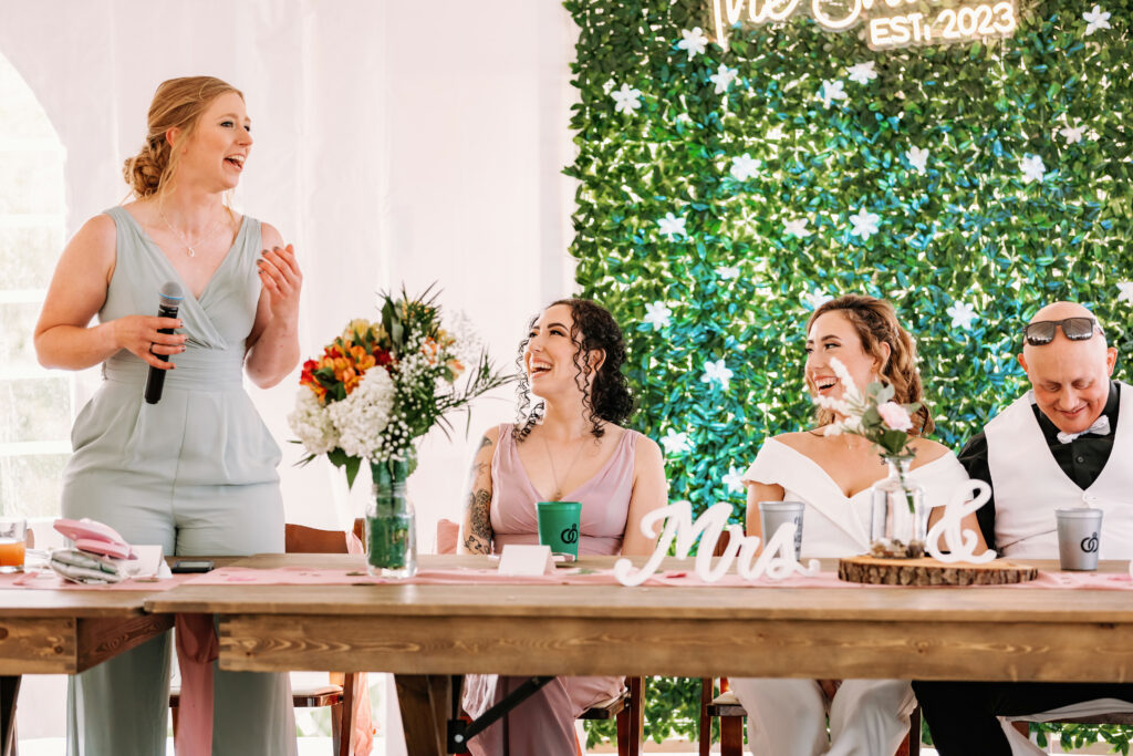 A person is giving a speech at a wedding reception, holding a microphone. The newlyweds and a guest are smiling at the table, with a "Mrs & Mrs" sign.