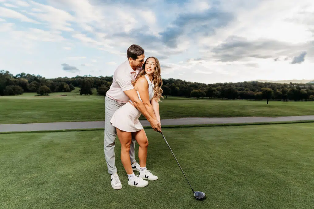 Two people on a golf course; one is teaching the other how to swing a golf club. Lush greenery and cloudy sky background.