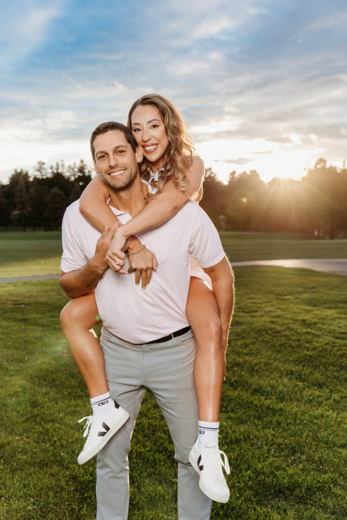 A person gives another person a piggyback ride on a grassy field at sunset, with trees and a cloudy sky in the background.