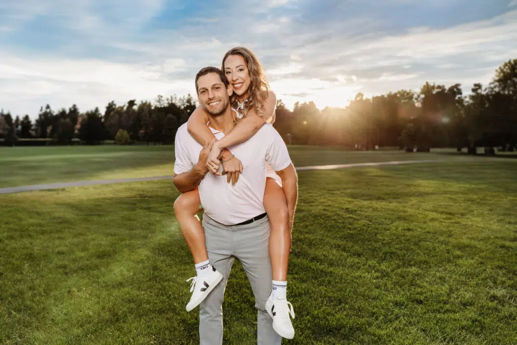 A person gives another a piggyback ride on a grassy field at sunset, surrounded by trees and a clear sky.
