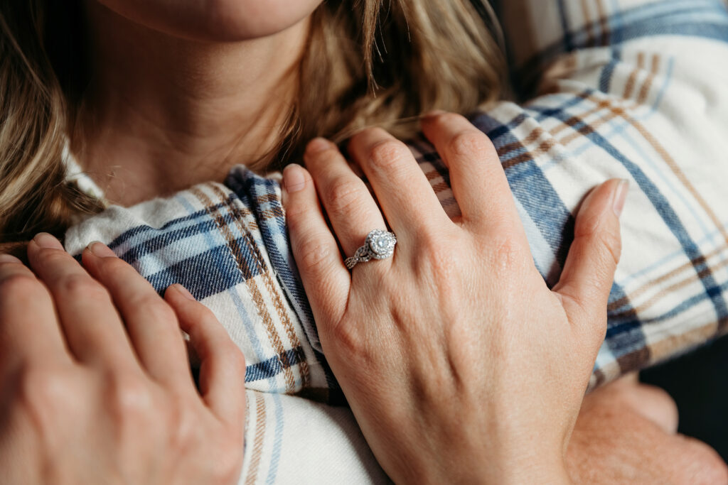 Close-up of a person's hand with an engagement ring on another person's shoulder, both wearing plaid shirts.