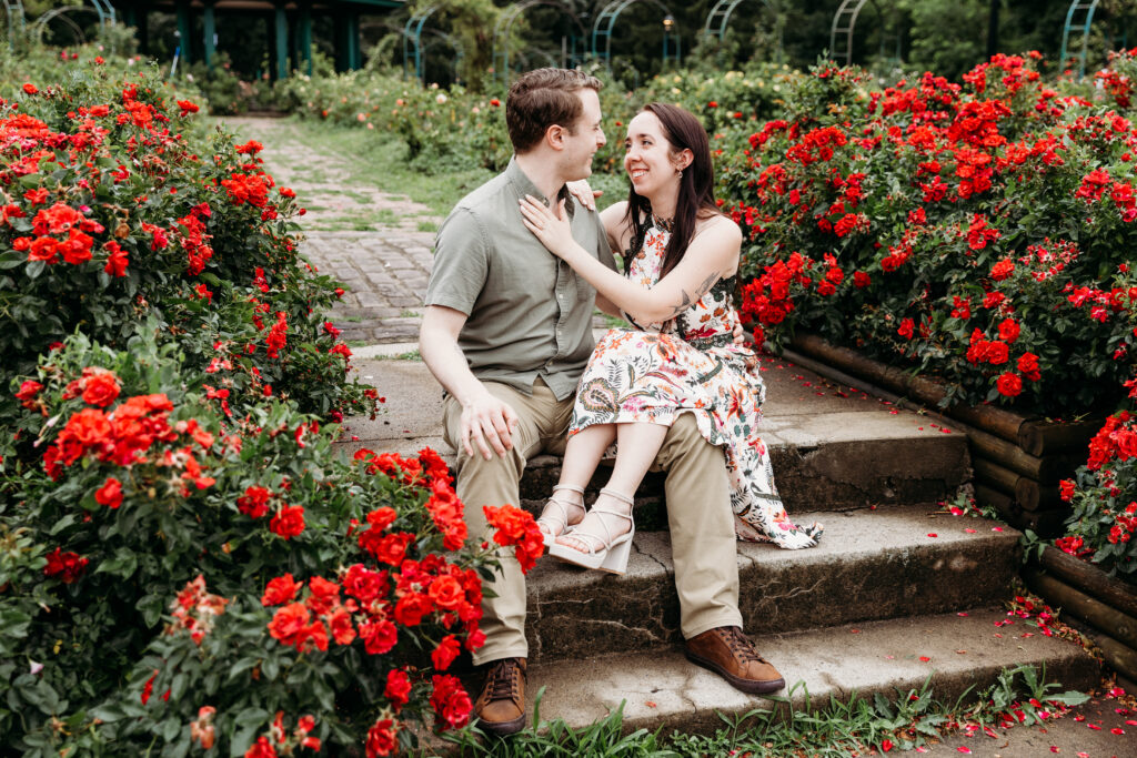 Two people sit on garden steps surrounded by vibrant red flowers, sharing a joyful moment in a lush, green park setting.