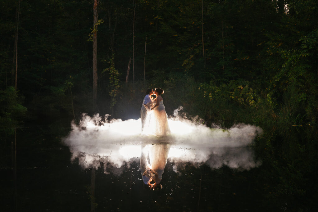 Two people embrace in a misty, wooded area, reflecting on a calm water surface; illuminated with dramatic lighting, surrounded by dense trees.