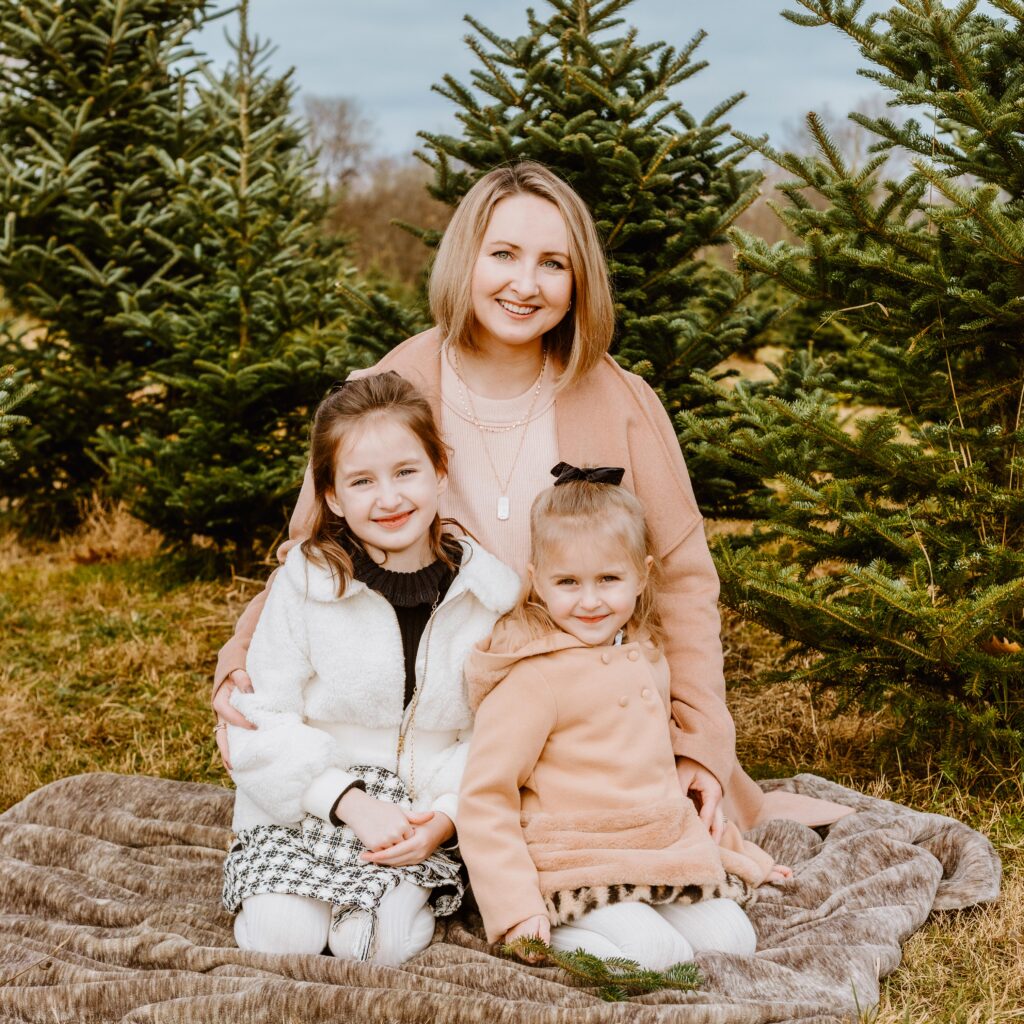 A smiling person with two young children, all wearing warm clothing, seated outdoors on a blanket with evergreen trees in the background.