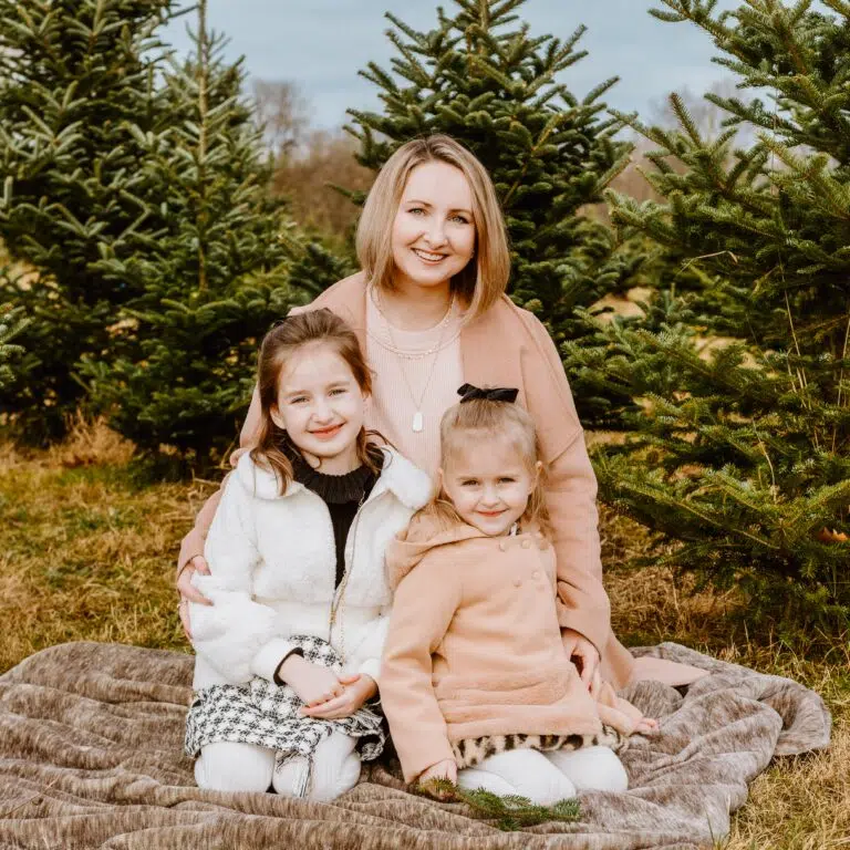 A smiling person with two young children, all wearing warm clothing, seated outdoors on a blanket with evergreen trees in the background.