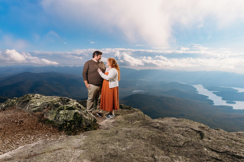Two people stand on a rocky mountain overlook, surrounded by expansive views of rolling hills and a distant lake under a partly cloudy sky.