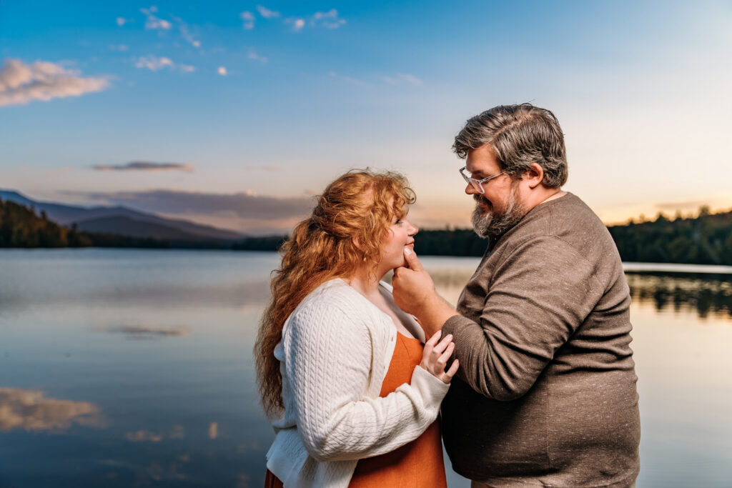Two people share a tender moment by a serene lake, with mountains and a clear sky in the background.