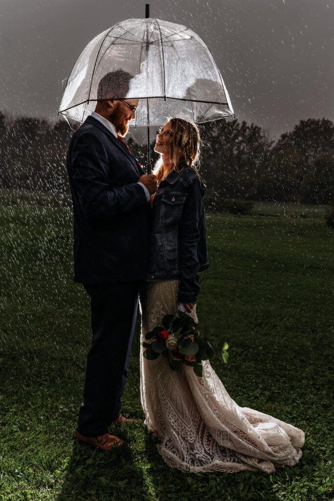 Two people stand under an umbrella in the rain, with greenery in the background. One holds a bouquet of flowers.