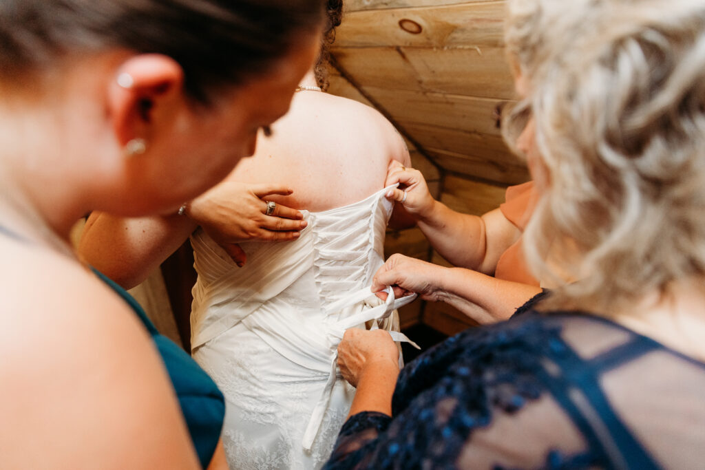 Three people help adjust the back of a wedding dress in a warmly lit wooden room, focusing on the intricate lacing details.
