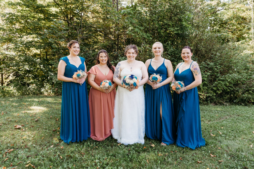Five people in formal dresses stand in a grassy area holding flowers, surrounded by lush green trees in the background.