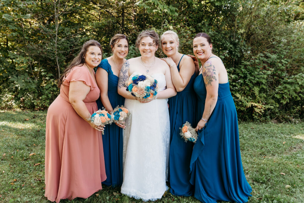 Five people in formal dresses stand smiling, holding bouquets, in a lush outdoor setting with trees in the background.