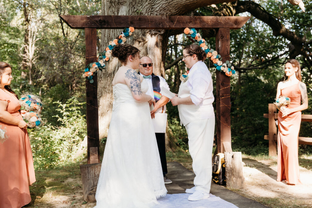 Two people exchange vows under a decorated wooden arch in a forest setting, with attendants holding bouquets and a person officiating.