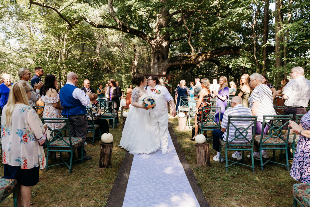 A wedding ceremony in a forest setting with a couple walking down an aisle surrounded by seated guests, sunlight filtering through trees.