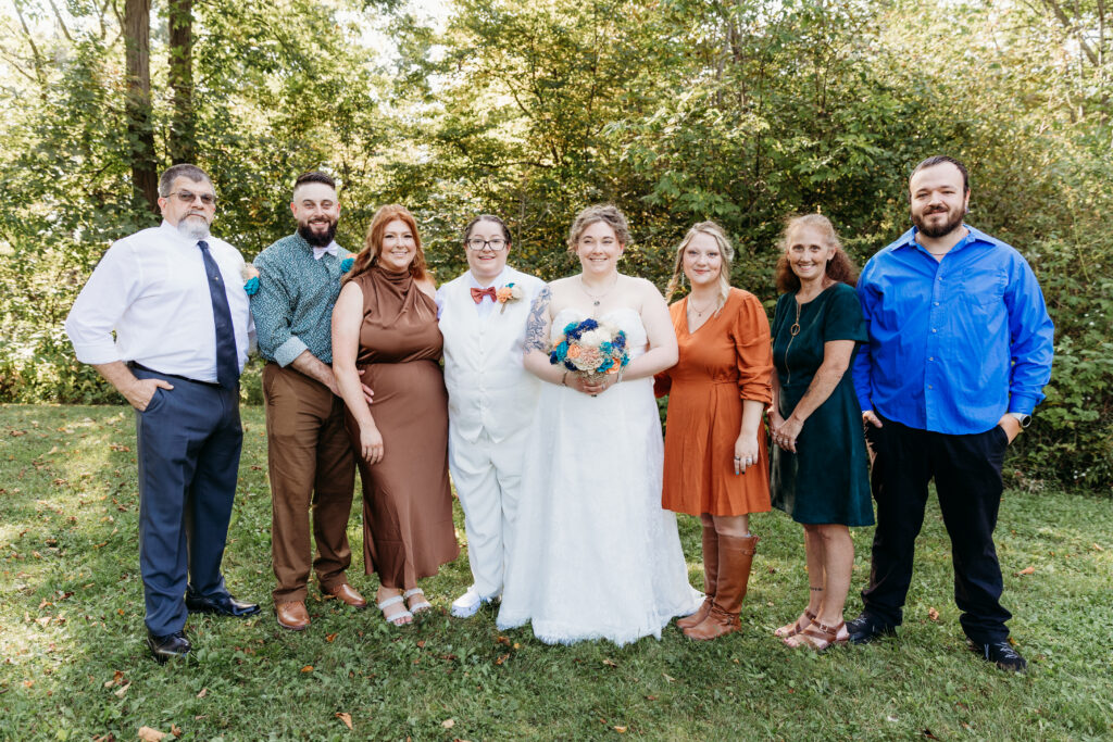 A group of eight people dressed in formal attire stand outdoors on grass, with trees in the background, appearing happy and relaxed.