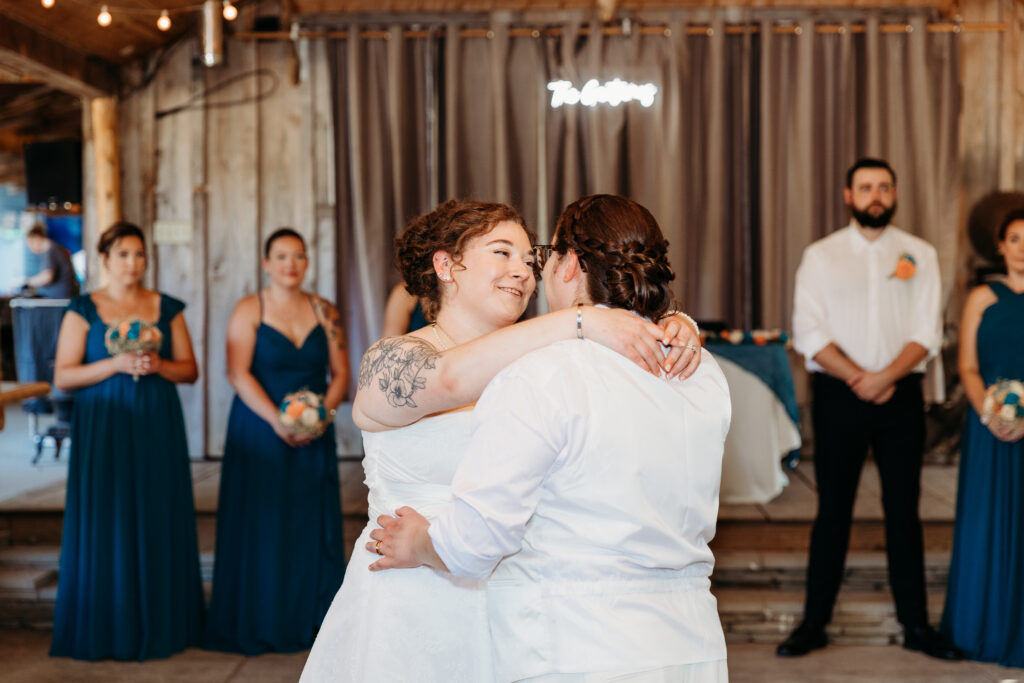 Two people dance at a wedding reception with onlookers dressed in blue. String lights hang overhead in a rustic venue setting.
