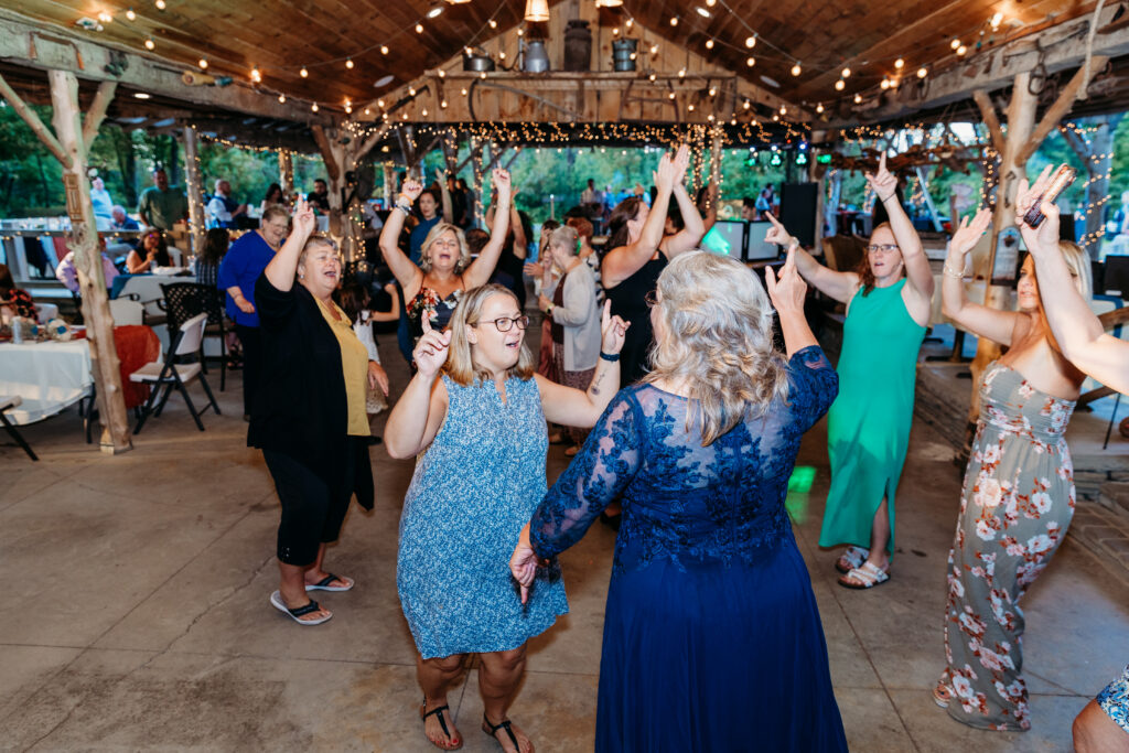A group of people happily dancing at a rustic venue adorned with string lights and wooden beams, creating a lively celebratory atmosphere.