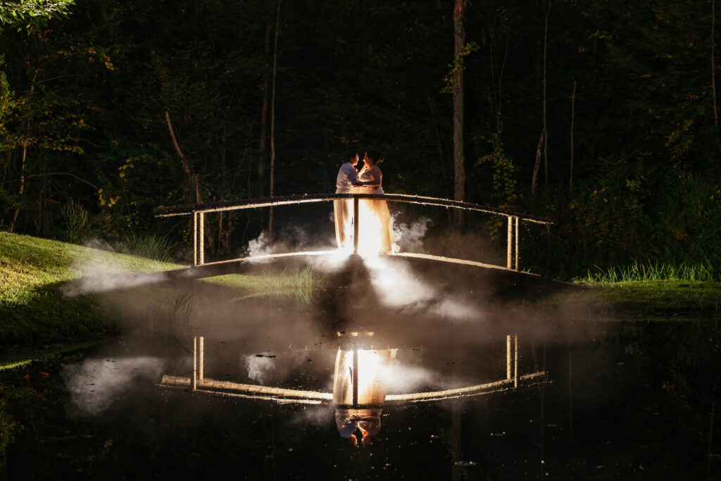 Two people stand on a wooden bridge over a misty pond, surrounded by trees at night, creating a romantic atmosphere.