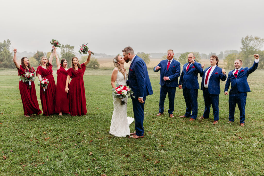 A wedding party poses outdoors, celebrating a couple's kiss. Six individuals in red dresses and blue suits cheer amid a grassy field.