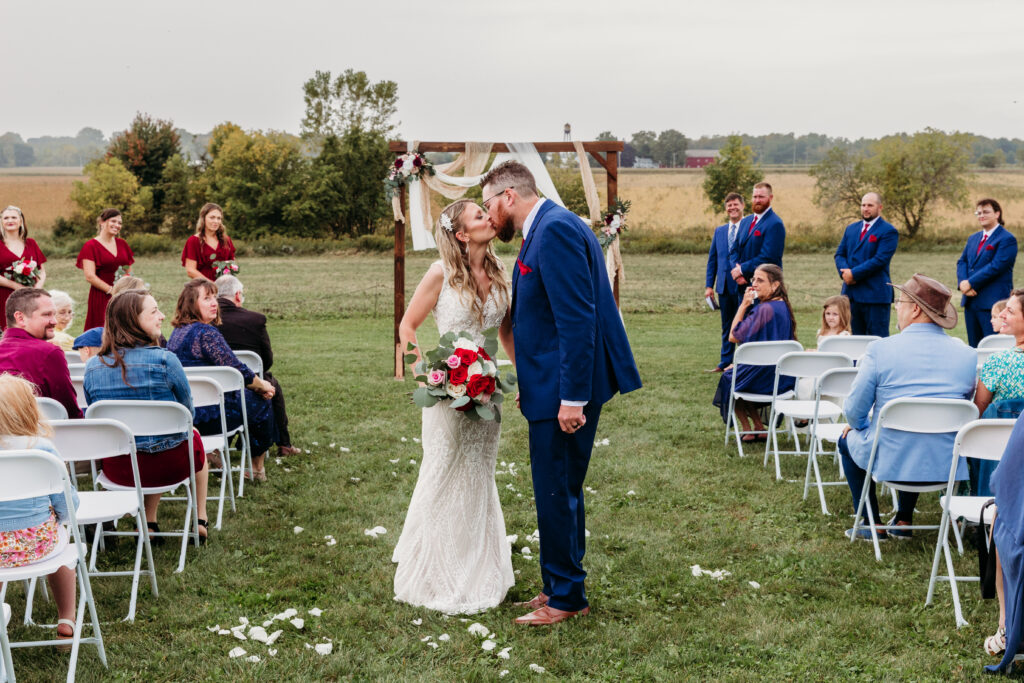A couple kisses under an arch at an outdoor wedding ceremony, surrounded by guests on white chairs in a scenic field setting.