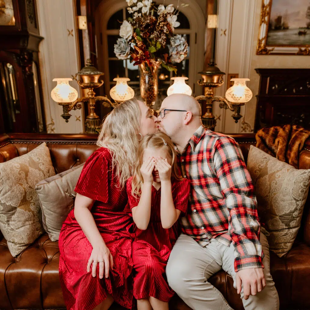 A child covers their eyes while two people kiss on a vintage sofa, surrounded by ornate decor and warm lighting.