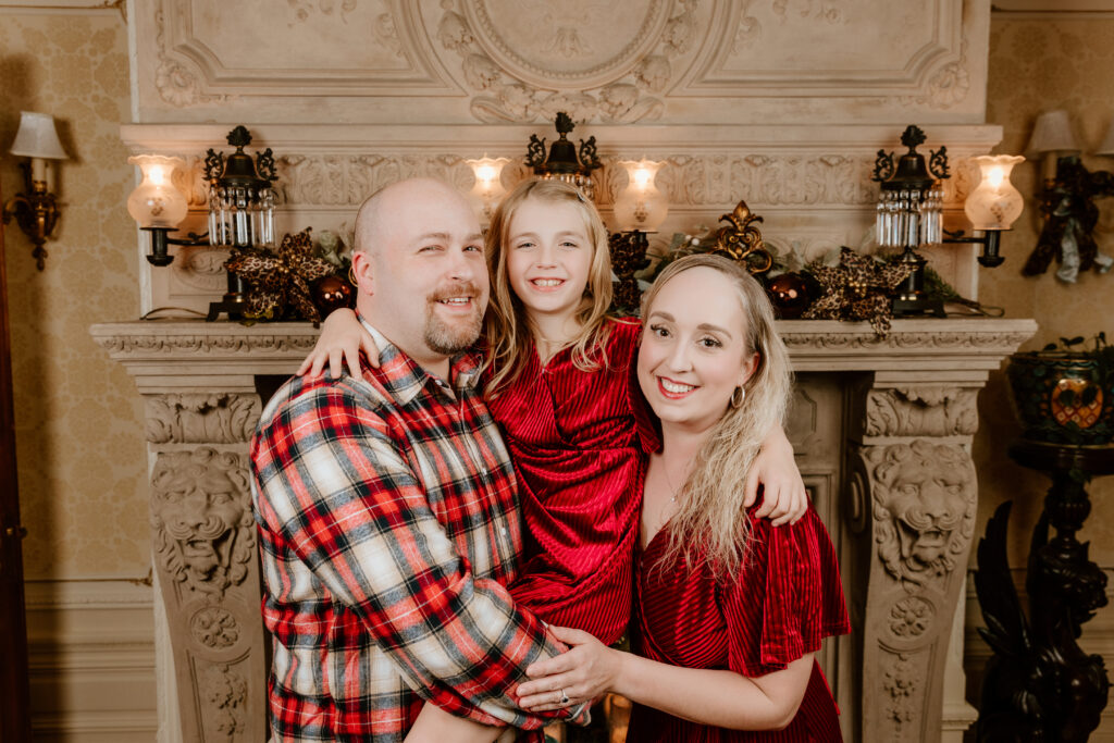 A smiling family portrait featuring three people standing in front of an ornate, decorated fireplace with festive ornaments and warm lighting.