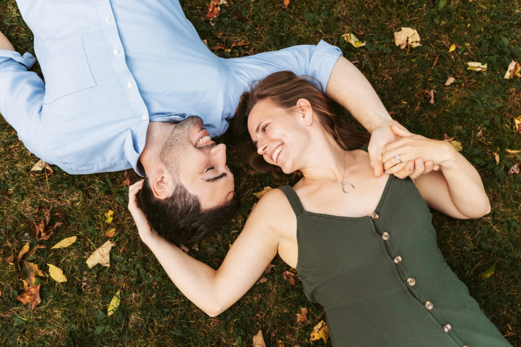 Two people lie on grass, holding hands and smiling warmly at each other, surrounded by scattered autumn leaves. Relaxed and joyful atmosphere.