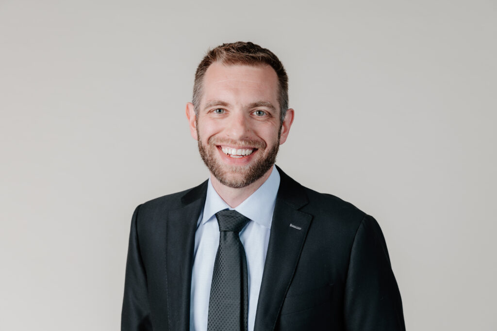 A person in a suit, smiling, against a plain background, with short hair and a beard. No landmarks or historical buildings present.