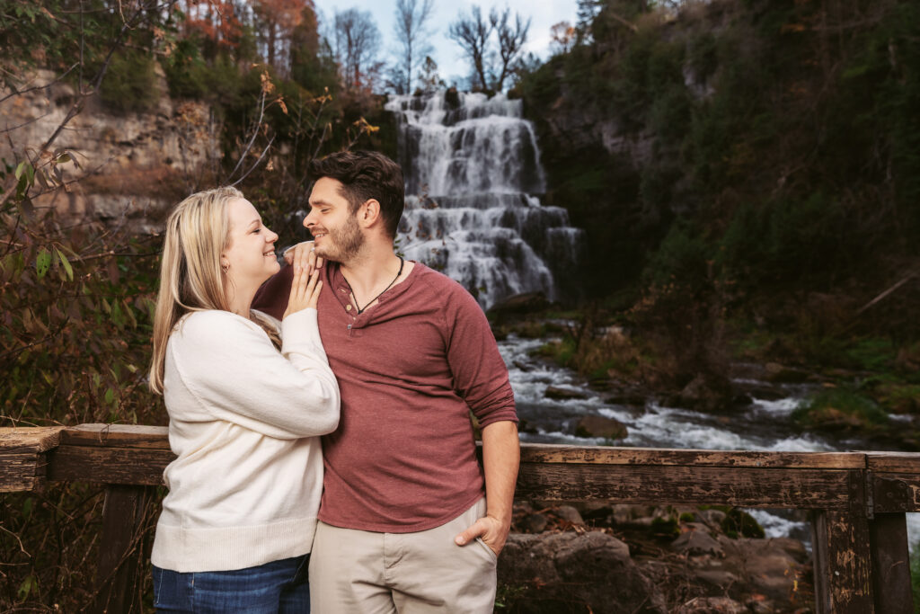 Two people stand on a wooden railing, smiling at each other, with a cascading waterfall and lush greenery in the background.